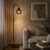 Brilliant lampa podłogowa Ligari, wygięte ramię, przydymiony klosz kroplowy, obok sofy
